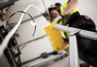 En sladd syns i bildens förgrund och i bildens bakgrund syns en elektriker som står på en stege