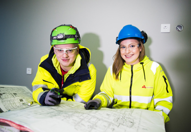 Två unga elektriker, iklädda varselkläder och hjälmar, som tittar in i kameran och ler medan en ritning ligger framför dem på ett bord