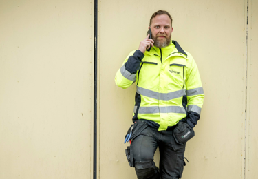 En person i varselkläder som står lutad mot en vägg med en telefon i handel