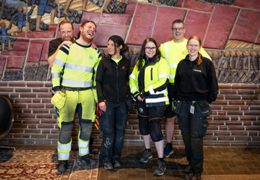 Två unga elektriker, iklädda varselkläder och hjälmar, som tittar in i kameran och ler medan en ritning ligger framför dem på ett bord
