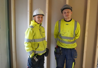 Två elektriker på en byggarbetsplats som tittar in i kameran och ler