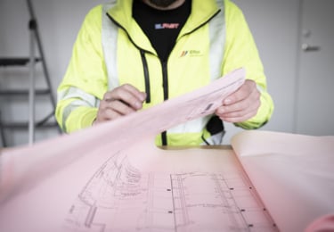 Elektriker i varseljacka som hanterar en stor ritning som ligger på ett bord